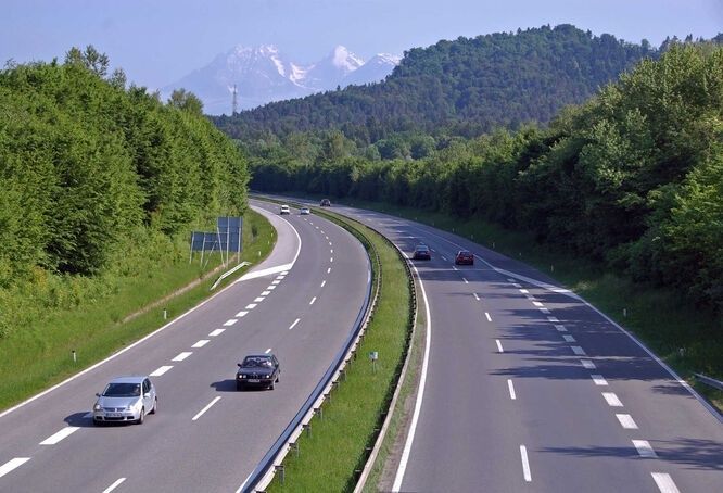 Bilar på en delad motorväg genom skog med alper i bakgrunden